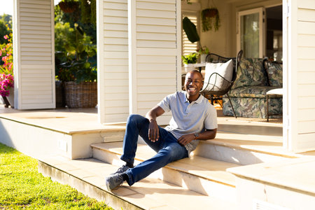 Outdoors, young African American man sitting on steps smiling at camera on sunny day. Lush greenery and cozy patio furniture in background creating warm and inviting atmosphere, unalteredの写真素材