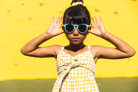 Biracial girl poses in playful sunglasses. She stands outdoors against a vibrant yellow background,...