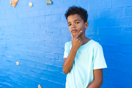 Biracial boy stands thoughtfully against a blue wall with copy space. His contemplative pose adds depth to the vibrant outdoor setting.の写真素材