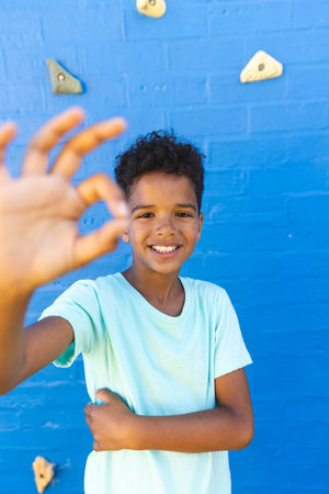 Biracial boy smiles in front of a climbing wall. His cheerful expression and the vibrant blue...