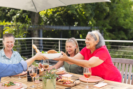 Diverse senior female friends sharing meal outdoors, passing dishes. Wearing casual summer attire, they are showing joyful expressions, unalteredの写真素材