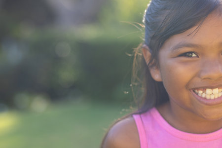 Outdoors, young biracial girl smiling brightly, wearing pink top, copy space. Greenery and sunlight background, creating warm and cheerful atmosphere, unalteredの写真素材