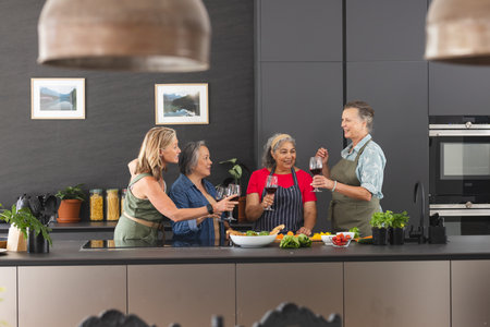 Diverse senior female friends holding wine glasses, laughing at home. Preparing food in modern kitchen, enjoying a lively atmosphere together, unalteredの写真素材