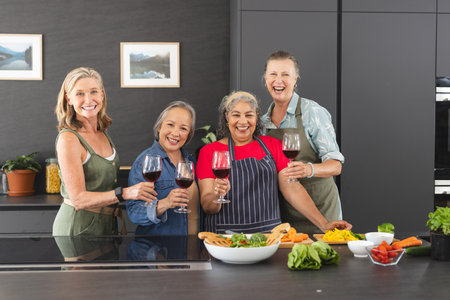Diverse senior female friends enjoying wine at home in kitchen. Sharing laughter, including Caucasian and Asian women, showcasing joyful expressions while holding glasses, unalteredの写真素材
