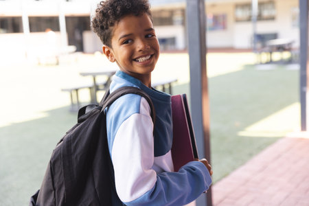 Biracial boy smiles at school, with copy space. He's ready for a new day of learning with his backpack and books.の写真素材