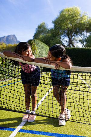 Outdoors, two biracial young sisters lean on a tennis net, smiling at each other. They are on outdoor tennis court with lush greenery and mountains in background, unalteredの写真素材