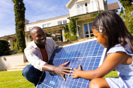 Outdoors, diverse father and daughter interacting with solar panel. They are in lush backyard with a large house in background, enjoying a sunny day, unalteredの写真素材