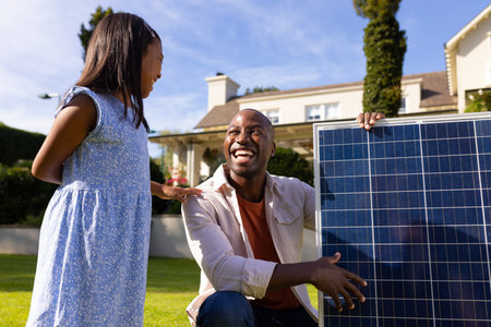 Outdoors, diverse father and daughter smiling at each other holding solar panel. They are outside in a sunny backyard with a large house in background, unalteredの写真素材