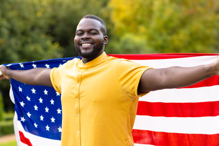Portrait of happy african american man covered with flag of usa with arms wide in garden. Independence day, fourth of july, patriotism, flags and lifestyle, unaltered.の写真素材