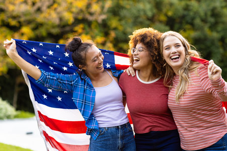 Happy diverse female friends covered with flag of usa in garden. Independence day, fourth of july, patriotism, friendship, flags and lifestyle, unaltered.の写真素材