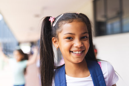 Biracial student smiling with long hair tied in pigtails. School hallway with blurred students in background, creating busy and lively atmosphere, unalteredの写真素材