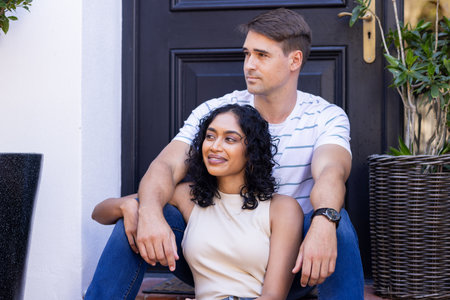Diverse couple sitting together on steps, looking content. Black background door with potted plants on either side, creating cozy atmosphere, unalteredの写真素材
