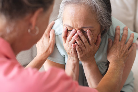 Asian senior woman covers her face as Caucasian senior woman comforts her. Retirement, friendship, senior living, happiness. Sitting on couch in cozy living room, sharing emotional moment, unalteredの写真素材