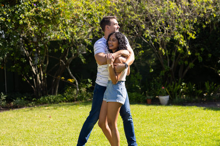 Young Caucasian man hugging young Asian woman from behind, both smiling and happy. They are standing in sunny garden with lush greenery and trees in background, unalteredの写真素材
