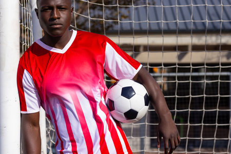 Young African American athlete holding soccer ball, leaning against goalpost. Outdoor sports field with net and building in background, green grass underfoot, and clear sky above, unalteredの写真素材