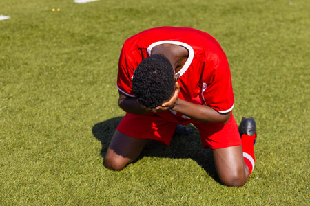 African American soccer player kneeling on grass field, looking down. Soccer field with white lines and green grass background in bright sunlight, unalteredの写真素材