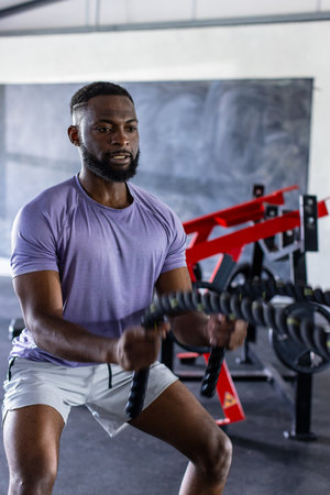 Exercising and working out, African American man using battle ropes in gym. Gym equipment and chalkboard background, creating intense and focused atmosphere, unalteredの写真素材