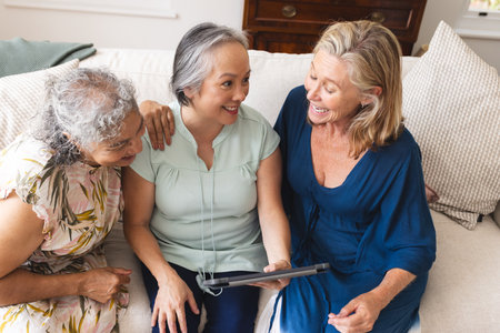 Diverse group of senior women friends using tablet at home on sofa. retirement, friendship, senior living, happiness, cozy living room with bookshelves and large windows, unalteredの写真素材