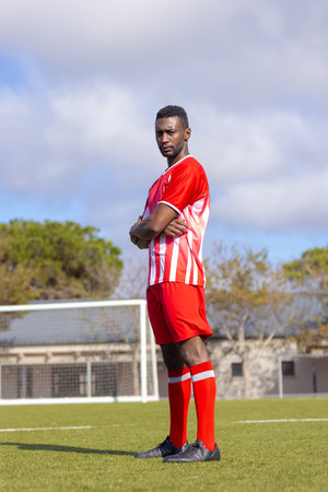 African American soccer player standing confidently on soccer field. Trees and buildings in background under a partly cloudy sky, creating a serene atmosphere, unalteredの写真素材