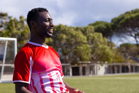 African American male fitness trainer wearing red sports jersey smiling outdoors, copy space. Soccer field with goalpost and trees in background, unalteredの写真素材