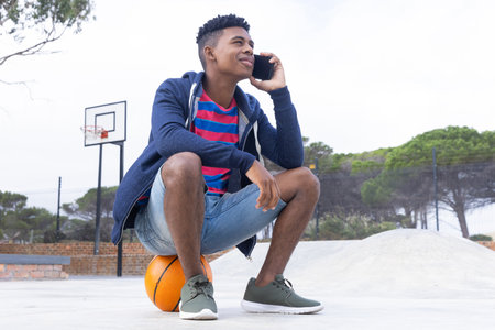 Sitting on basketball, African American teenage boy talking on smartphone outdoors. Sports, communication, urban, leisure, technology, youth, unalteredの写真素材