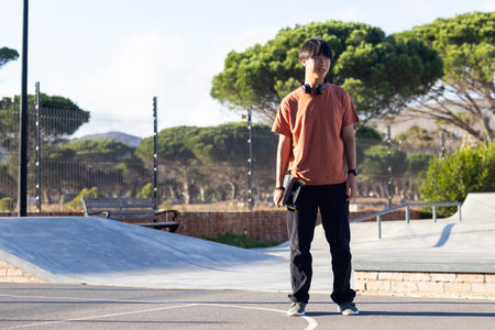 Teenage boy with headphones standing in skate park, enjoying outdoor hangout, copy space. Youth, music, lifestyle, skateboarding, casual, fun, leisure, unalteredの写真素材