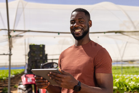 African American farmer using tablet for managing hydroponic farm, smiling outdoors. Agriculture, farming, technology, organic, produce, sustainable, unalteredの写真素材