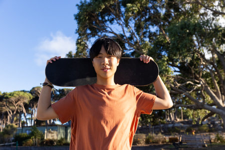 Asian teenage boy holding skateboard behind head, smiling outdoors in park. Youth, skateboarding, lifestyle, fun, leisure, urban, unalteredの写真素材