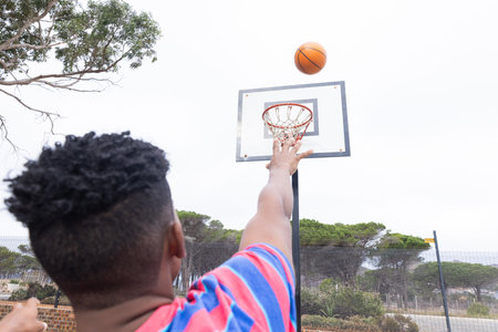 Playing basketball, male student shooting ball towards hoop in outdoor court. Sports, athletics, competition, teamwork, unalteredの写真素材
