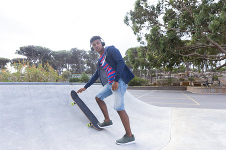 Skateboarding in park, African American teenage boy wearing headphones enjoying day. Sports, urban, leisure, music, outdoors, active, unalteredの写真素材