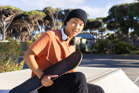 Teenage boy holding skateboard and wearing headphones, relaxing at skate park. Youth, leisure, skateboarding, urban, lifestyle, music, unalteredの写真素材