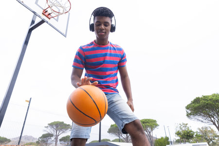 Dribbling basketball, teenage boy wearing headphones enjoying outdoor activity on court. Sports, active, youth, enjoyment, unalteredの写真素材
