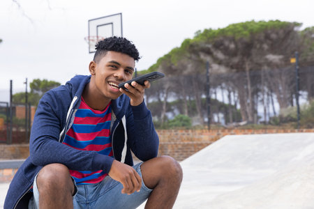 African American teenage boy using smartphone voice command, sitting at skatepark, copy space. Youth, technology, communication, leisure, summer, lifestyle, unalteredの写真素材