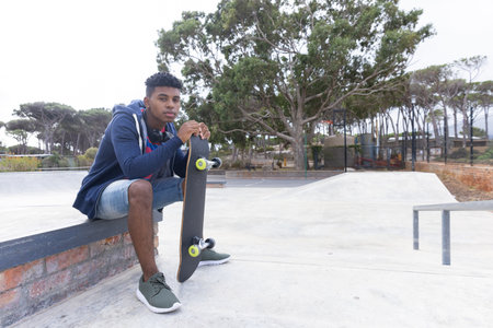 Sitting on bench, teenage boy holding skateboard, relaxing at skate park, copy space. Youth, recreation, skateboarding, outdoor, lifestyle, leisure, unalteredの写真素材