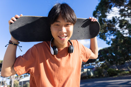 Holding skateboard and wearing headphones, teenage boy smiling outdoors. Youth, lifestyle, urban, leisure, music, trendy, unalteredの写真素材