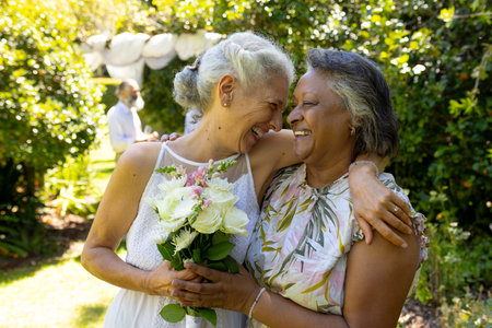 Senior women hugging and smiling, holding bouquet of flowers in garden. Friendship, outdoors, celebration, joy, bonding, happiness, elderly, unalteredの写真素材