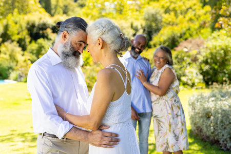 Dancing together, senior bride and groom enjoying outdoor celebration with friends nearby smiling. Seniors, outdoors, friendship, happiness, enjoyment, lifestyleの写真素材