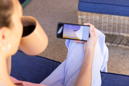 Holding coffee cup, woman browsing travel website on smartphone at home. Technology, lifestyle,...