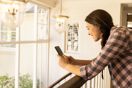 Using smartphone, young woman leaning on railing and smiling at home, copy space. Technology, lifestyle, casual, relaxation, happy, indoorsの写真素材