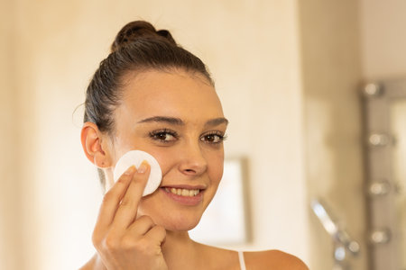 Smiling woman applying facial cleanser with cotton pad in bathroom. Skincare, beauty, hygiene, wellness, self-care, cosmeticsの写真素材
