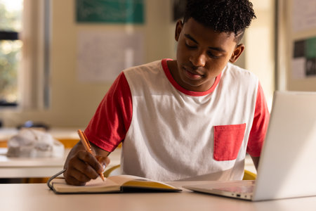 In high school, teenage boy writing in notebook while using laptop in classroom. Education, studying, learning, technology, notes, teachingの写真素材