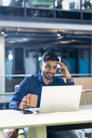Smiling businessman holding coffee cup and using laptop in modern office, copy space. professional,...