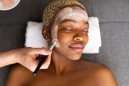 Hand of beautician giving facial to smiling african american woman with brush. Spa, relaxation,...