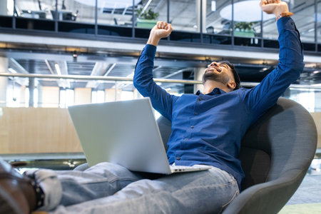 Celebrating success, man with laptop sitting in modern office chair. achievement, business, professional, celebration, productivity, technologyの写真素材