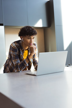 Using laptop, teenage boy with earbuds focusing on work in modern kitchen, copy space. Technology,...