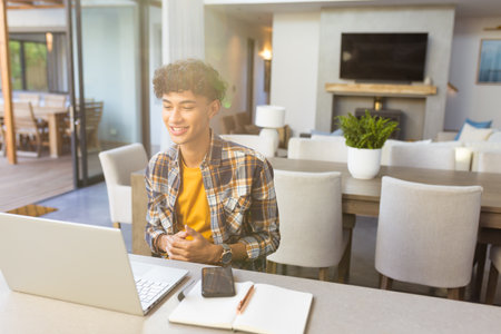 Smiling teenage boy using laptop and taking notes at home office desk, copy space. Remote working,...