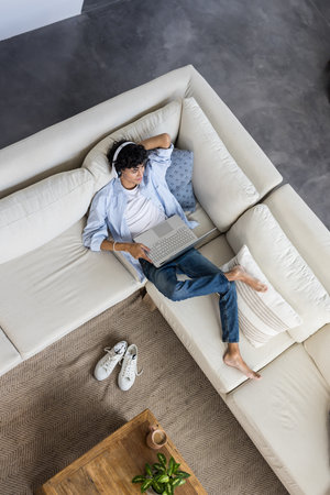 Relaxing on sofa, young man using laptop and wearing headphones at home. Remote working, relaxation, technology, home office, digital nomad, casualの写真素材