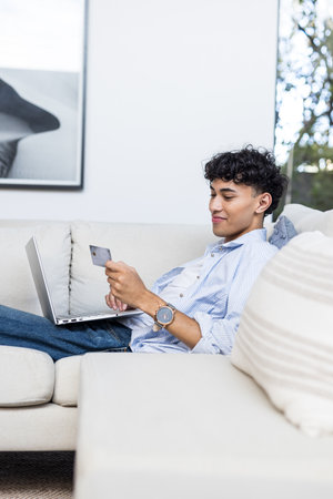 Using credit card and laptop, young man shopping online while relaxing on couch. E-commerce, online shopping, technology, internet, digital, convenienceの写真素材