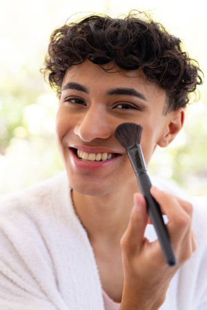 Smiling young man applying makeup with brush, wearing white robe at home. Beauty, cosmetics, skincare, self-care, grooming, facialの写真素材