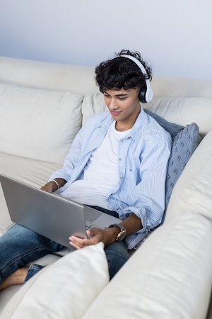 Using laptop and wearing headphones, teenage boy relaxing on couch at home. Technology, relaxation,...